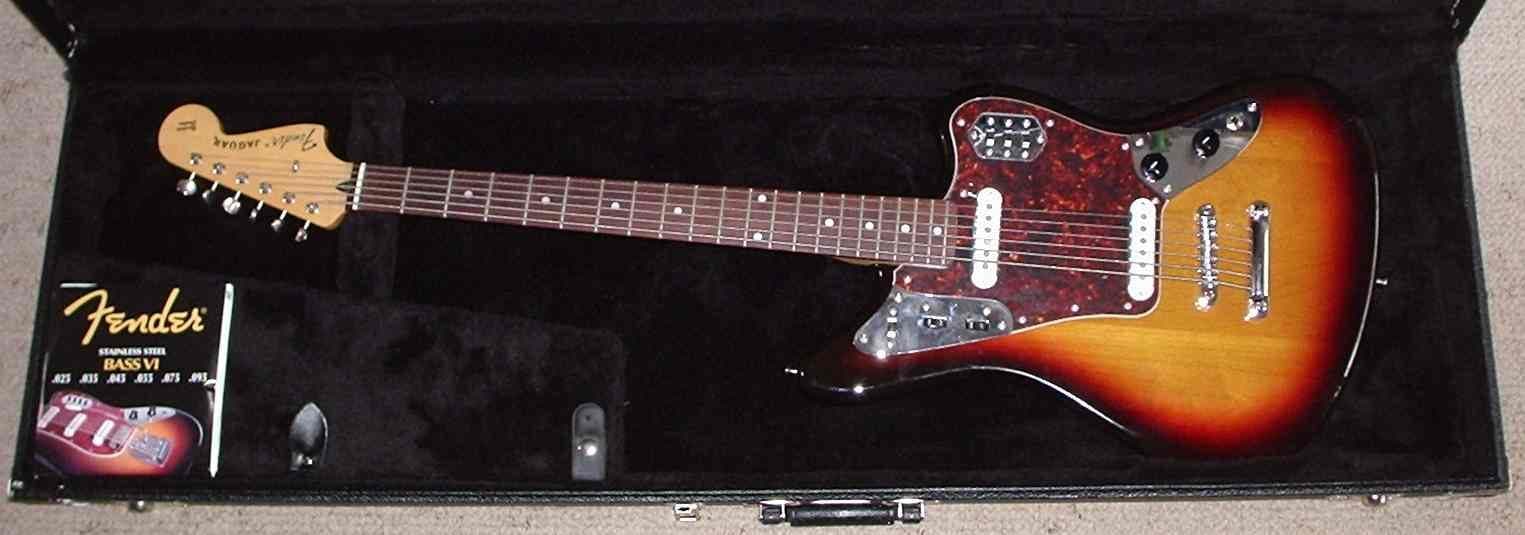 A long-scale Fender baritone-range guitar in its case beside a pack of strings.