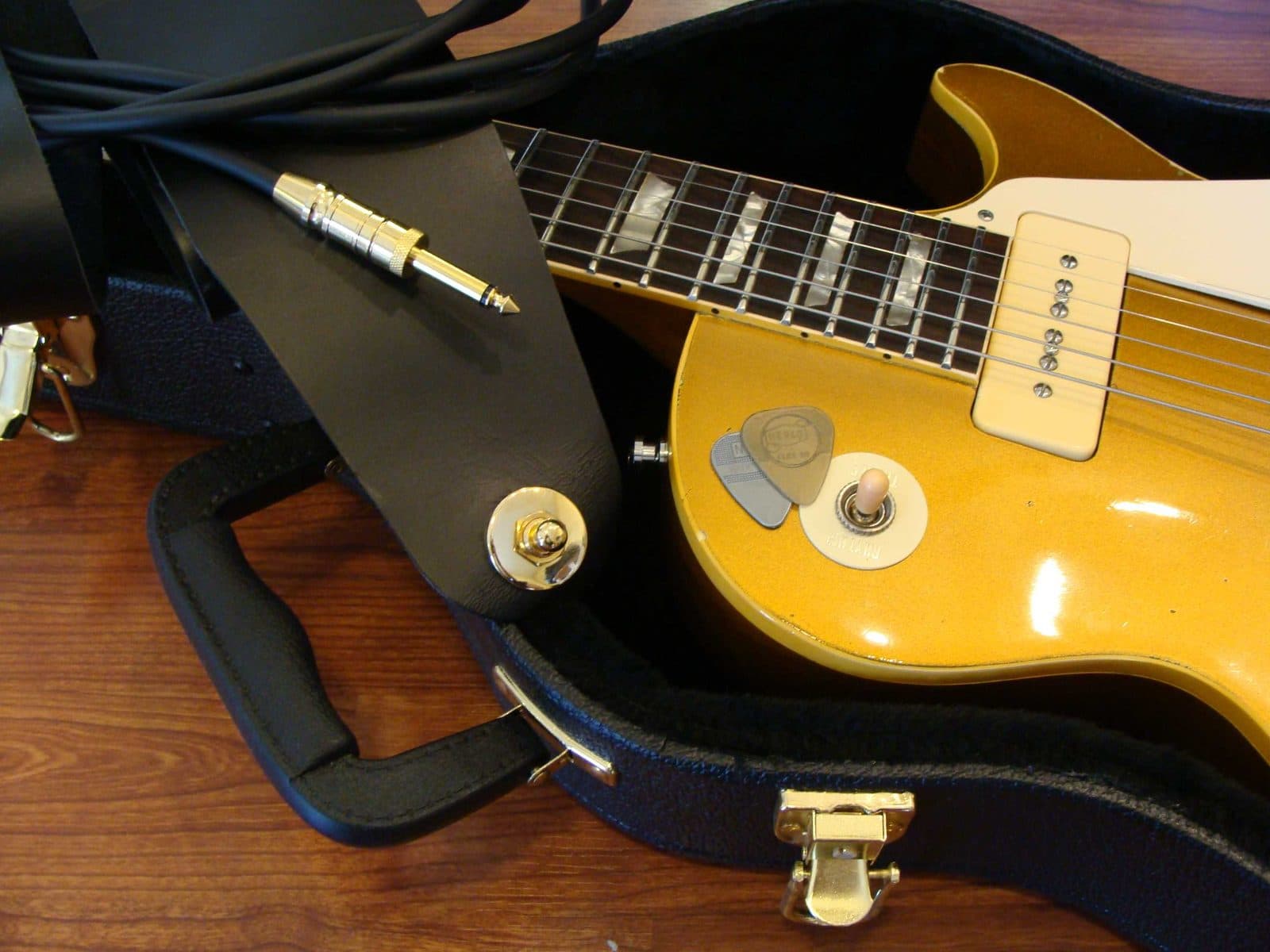 A gold-top electric guitar in its case with a cable and picks.