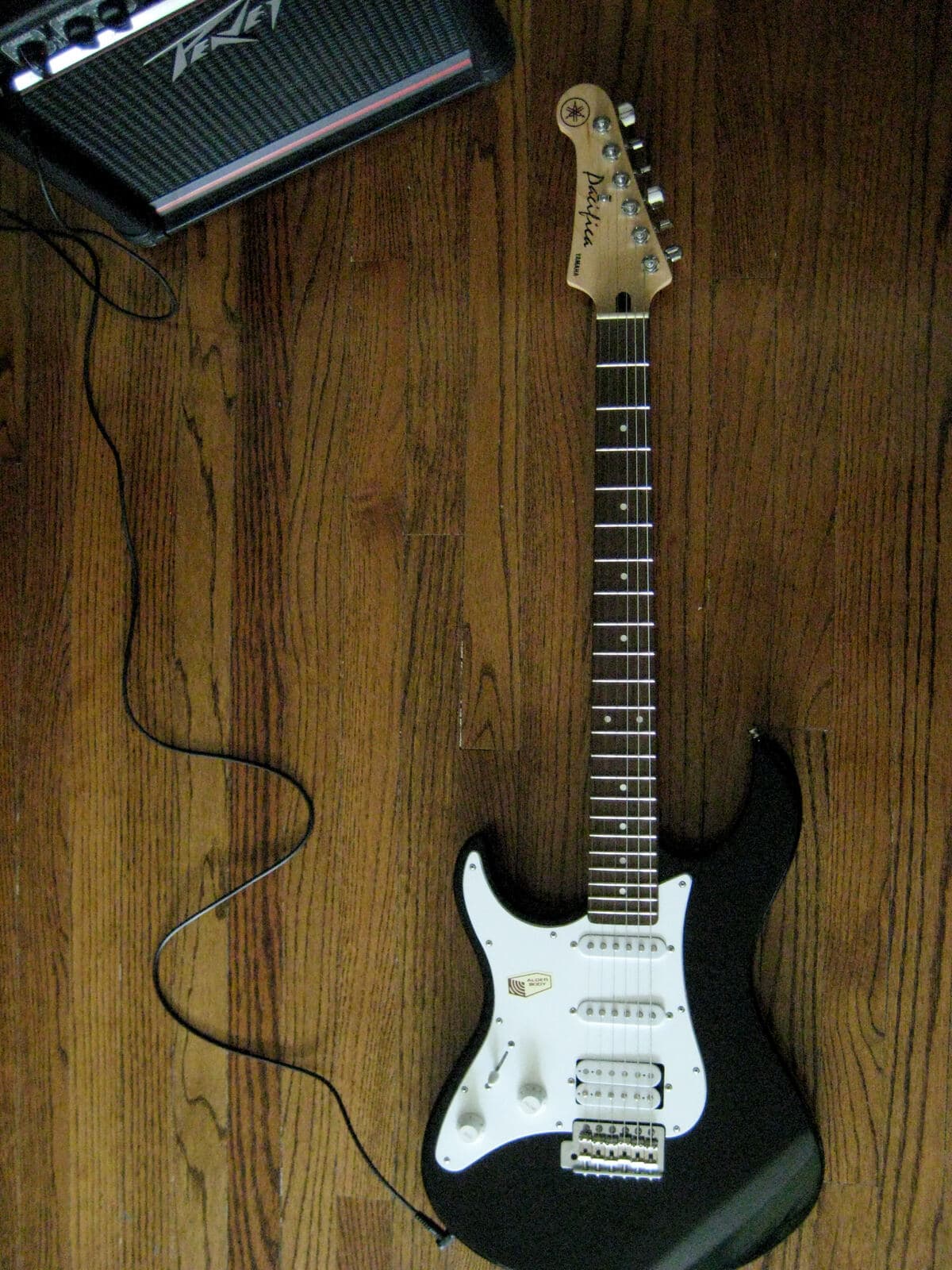 A black Yamaha Pacifica electric guitar with HSS pickups and the Pacifica logo on its headstock, resting on a wood floor next to a Peavey amplifier